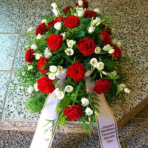 Bouquet of red roses and white flowers with greenery and two white ribbons bearing names, placed on a speckled tile floor.