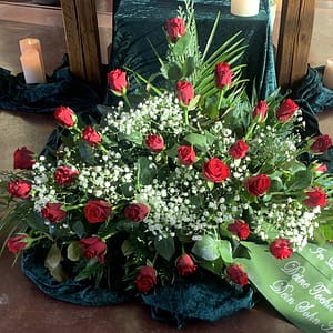 Funeral casket spray with red roses, white baby’s breath, and greenery on dark velvet with surrounding candles.