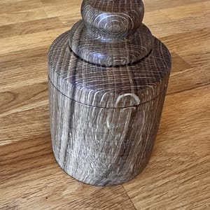 Close-up of a carved dark wooden container with a rounded knob on top, sitting on a wooden floor.